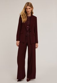 Burgundowy garnitur składający się z dopasowanej marynarki i spodni z szerokimi nogawkami. Teksturowana tkanina, detale z guzikami na górze oraz buty z szpiczastym noskiem.