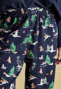 Tumed pajama püksid pehmest kangast, millel on jõuluteema print, kus on kujutatud lumeinimesi, puid ja pidulikke figuure kogu pinnal.