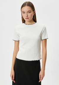 Hvid ribbet T-shirt med korte ærmer, der har en bølget sort kant rundt om udskæringen og ærmerne, samt en let cropped kant.