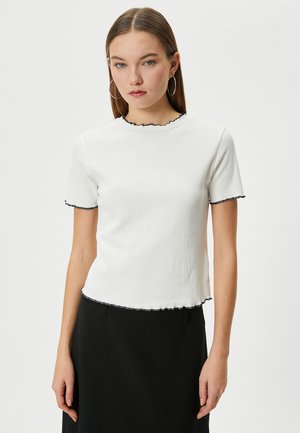 Camiseta blanca de rayas con mangas cortas, que presenta un borde negro ondulado alrededor del escote y las mangas, y un dobladillo ligeramente recortado.