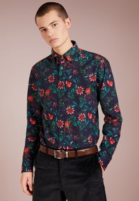 Blumenhemd aus Baumwolle mit einem dunkelblauen Hintergrund, lebhaften roten Blumen, grünen Blättern und einem Button-Down-Kragen. Kombiniert mit schwarzen Hosen.
