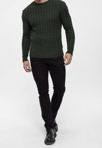 Mörkgrön kabelstickad tröja med rund halsringning, kombinerad med svarta slim-fit jeans och svarta snörskor.