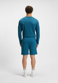 Teal langemouwen top en bijpassende shorts, gemaakt van textuurstof. Heeft een ronde halslijn en een elastische tailleband. Witte sokken zichtbaar.