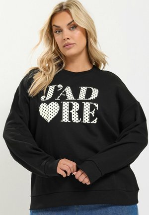 Lyshåret kvinde iført en sort sweatshirt med hvide polkaprikkede bogstaver, der staver "J'ADORE", og et hjertesymbol, poserer mod en ensfarvet baggrund.
