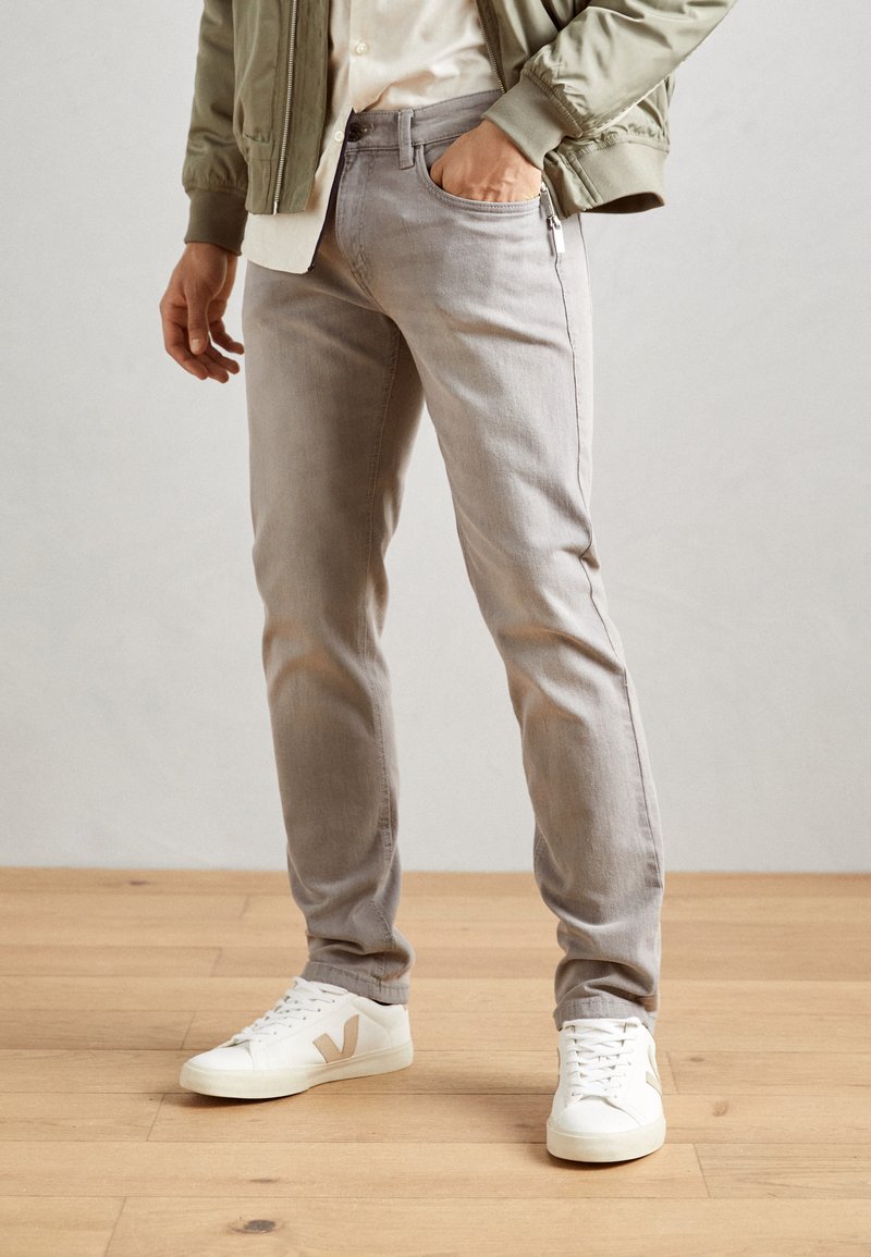 Light gray slim-fit jeans in denim, featuring a front pocket, subtle whiskering, worn with white sneakers and an olive green bomber jacket.