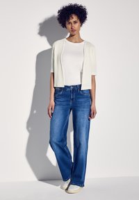 Vit kortärmad cardigan över en enkel vit skjorta, tillsammans med blå vida jeans och vita sneakers, mot en neutral bakgrund.