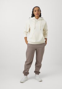 Felpa crema con cappuccio e lacci, tasca frontale; abbinata a pantaloni jogger marroni con gambe affusolate e polsini elasticizzati; sneakers bianche.
