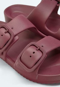 Gros plan sur des sandales en caoutchouc bordeaux avec deux brides à boucles réglables et des semelles texturées.