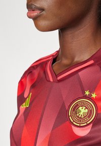 Raudonas ir tamsiai raudonas futbolo marškinėlis su V formos iškirpimu, žalia siuvinėta logo ir dinamiškais raštais su raudonos spalvos atspalviais.