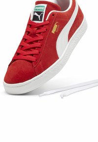 Røde ruskinds sneakers med hvid gummisål, prydet med røde snørebånd, et grønt mærke og en hvid sidestribe; inkluderer ekstra hvide snørebånd.