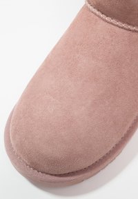 Primer plano de la punta de una bota de ante rosa polvoriento con costuras visibles y borde de suela texturizado sobre un fondo blanco.