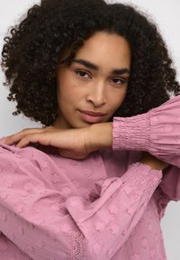 Blouse rose à texture avec des manches froncées, présentant des motifs floraux et des accents en dentelle. Tissu doux, mettant en avant une coupe décontractée et un drapé léger.