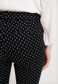 Svarta mönstrade byxor med en vit diamantdesign, med en bakficka och en mjuk textur. Synlig midja och en vit blus i närheten.