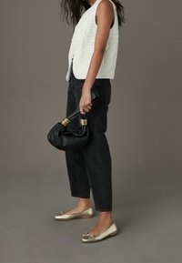 Weißes gesteppte Weste, schwarze hochtaillierte Jeans und goldene Ballettschuhe mit Schleifendetail. Schwarze Handtasche mit goldenen Akzenten, in der Hand gehalten.