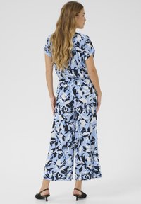 Jumpsuit con motivo floreale nei toni del blu chiaro, nero e bianco; vestibilità ampia con maniche corte e cintura da annodare in vita. Calzature con tacco nero.
