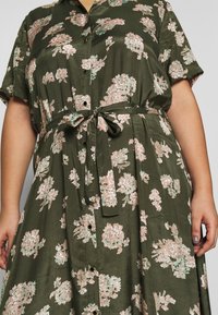 Robe florale verte avec des manches courtes, boutonnière à l'avant et ceinture à la taille. Présente des motifs de fleurs roses et blanches sur un tissu lisse.