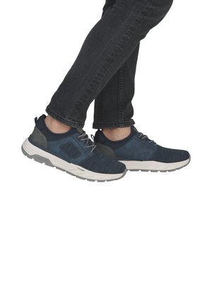 Personne portant un jean foncé et des baskets de sport bleu marine avec des semelles blanches, debout avec un pied légèrement levé.