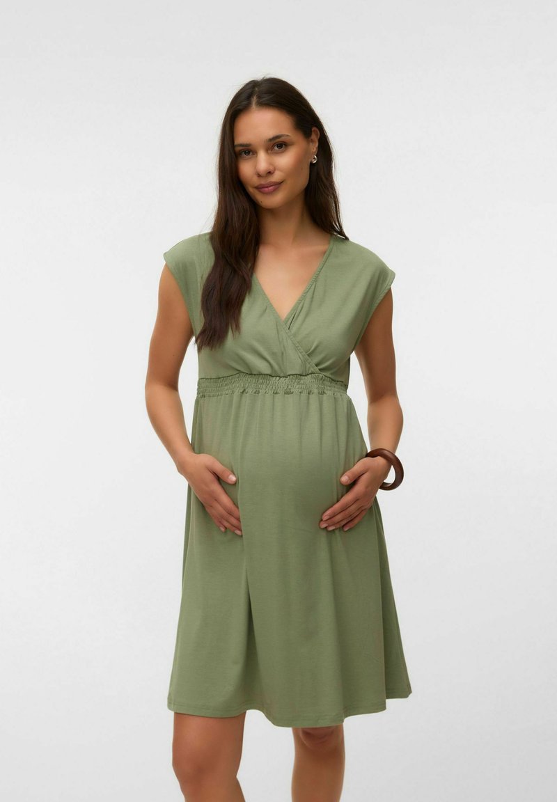 Femme enceinte aux longs cheveux foncés portant une robe courte à manches courtes couleur vert olive, tenant doucement son ventre, debout devant un fond uni.