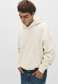 Hellbeige Kapuzenpullover aus weichem Stoff, mit einer Kängurutasche und gerippten Bündchen. Kombiniert mit lässigen blauen Jeans im entspannte Schnitt.