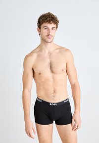 Schwarze Boxer-Brief mit einem weißen "BOSS"-Logo am Bund; glatt gewebter Stoff und taillierte Passform, getragen von einem männlichen Modell, das aufrecht steht.