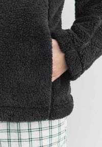 Zwarte fleece jas met een textuur en een fluffy afwerking. Kenmerkt zich door een afgeronde zoom en elastische manchetten, met een hand in de zijzak.