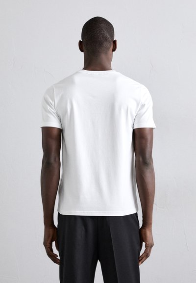 T-shirt bianca in cotone con maniche corte, scollo rotondo e vestibilità rilassata. Indossata con pantaloni scuri, mostrando un design semplice.