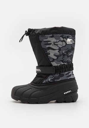 Bota de invierno de camuflaje negro y gris con suela gruesa, correa ajustable, parte superior elástica con cierre de ajuste, y logo "Sorel" en el lateral y la correa.