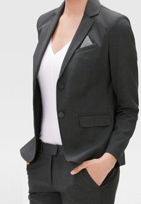 Blazer su misura grigio scuro con rever a gola a lancia, due bottoni e una tasca sul petto con un fazzoletto grigio a pois. Abbinato a una camicia bianca.