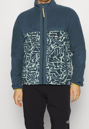 Mann trägt blaue Fleecejacke mit abstraktem hellblauem und marinefarbenem Muster, Reißverschluss vorne, und schwarze Hose vor einfarbigem Hintergrund.