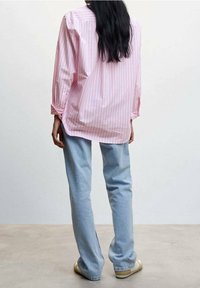 Personne aux longs cheveux foncés portant une chemise oversize rayée rose, un jean bleu clair et des chaussures beige, debout de dos sur un sol uni.