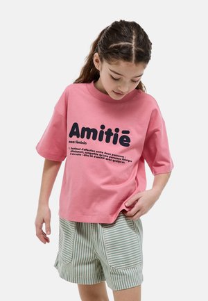 Fille aux cheveux tressés portant un t-shirt rose "Amitié" et un short rayé vert, regardant vers le bas, debout devant un fond uni.
