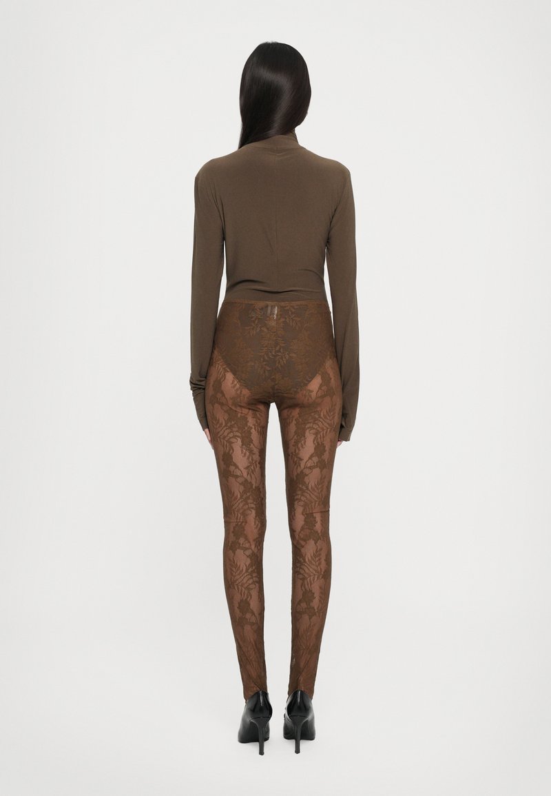 Braunes langärmeliges Oberteil, kombiniert mit durchsichtigen braunen Leggings mit floralem Muster, getragen mit schwarzen High Heels, von hinten betrachtet.