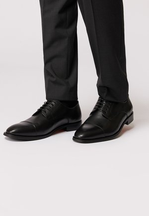 Schwarze Lederschuhe mit runden Zehen, Kapuze-Design, tonal abgesteppt und Schnürverschluss. Getragen zu maßgeschneiderten dunklen Hosen.