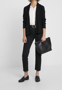 Černý žebrovaný cardigan přes bílou košili s knoflíky, doplněný černými džíny a námořnickými mokasíny. Doplňky zahrnují černou kabelku a zlatou sponu opasku.