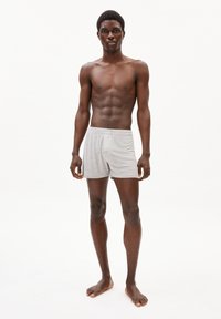 Un slip boxeur en coton gris porté par un modèle masculin torse nu avec une silhouette élancée, standant pieds nus sur un fond blanc uni.