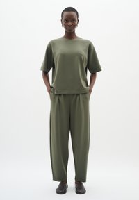 Žena stojí na bílém pozadí, oblečená v olivově zeleném volném tričku s krátkým rukávem a odpovídajících širokých kalhotách, s černými nazouvacími botami.