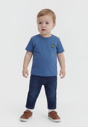 Tout-petit debout portant une chemise à manches courtes bleue avec un logo jaune, un jean bleu foncé, des chaussettes blanches et des baskets marron sur un fond uni.