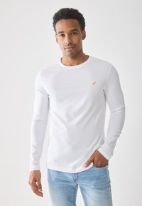 Hvit langermet T-skjorte laget av myk stoff, med en liten gul logo på brystet. Kombinert med lyseblå jeans.