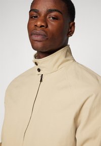 Chaqueta beige con cuello alto, que cuenta con un cierre de cremallera y dos botones marrones, fabricada con una tela lisa y estructurada.