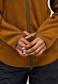 Giacca in pile marrone con superficie testurizzata e polsini aderenti, abbinata a pantaloni trapuntati blu navy. Chiusura a zip frontale con dettagli di rifinitura a contrasto.