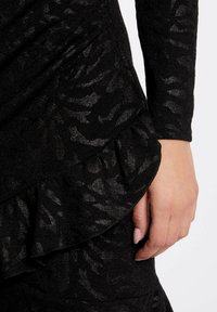 Robe noire texturée avec un ourlet froncé, présentant un motif subtil et des manches longues. Le matériau semble léger et légèrement brillant.