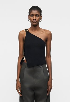 Femme portant un haut noir à une épaule orné de deux grands anneaux dorés et un pantalon taille haute sur mesure sombre, debout devant un fond uni.