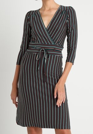 Femme portant une robe portefeuille ajustée longueur genoux avec des rayures verticales noires, blanches, rouges et vertes et des manches trois-quarts.
