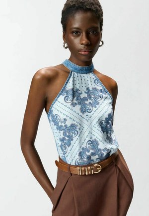 Donna che indossa un top senza maniche blu con fantasia paisley infilato in pantaloni marroni a vita alta con cintura color tabacco, in piedi con le mani in tasca.