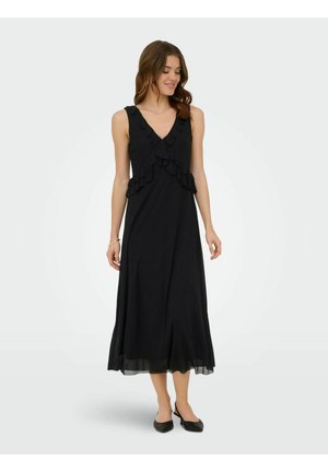 Femme portant une robe midi noire sans manches avec des détails volantés, debout et regardant vers le bas avec un sourire doux sur un fond uni.