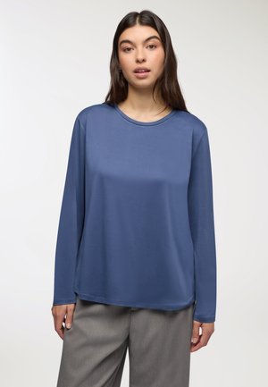 Camicia a maniche lunghe di colore blu scuro, realizzata in tessuto liscio, con colletto rotondo e vestibilità comoda, abbinata a un paio di pantaloni chiari grigi con texture.