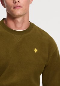 Maslinasto zelena dukserica s mekanom teksturom i rebrastim izrezom. Ima mali žuti izvezeni logo na prsima.