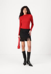 Rode geribde lange mouwen top, zwarte franje mini rok, zwarte knielange laarzen en een kleine rode handtas. Eenvoudige achtergrond.