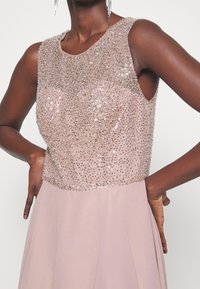 Klänning med paljettdekoration, rundad urringning och ärmlös design. Mjuk, ljusrosa tyg med en flödande kjol och detaljerad textur.