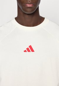 Biała bawełniana koszulka z okrągłym dekoltem i krótkimi raglanowymi rękawami. Na przodzie znajduje się czerwone logo Adidas w postaci trzech ukośnych pasków.